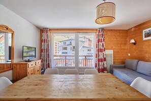 Une pièce comprenant une table à manger en bois, des chaises blanches, un téléviseur posé sur un meuble en bois, un canapé et une fenêtre avec des rideaux.