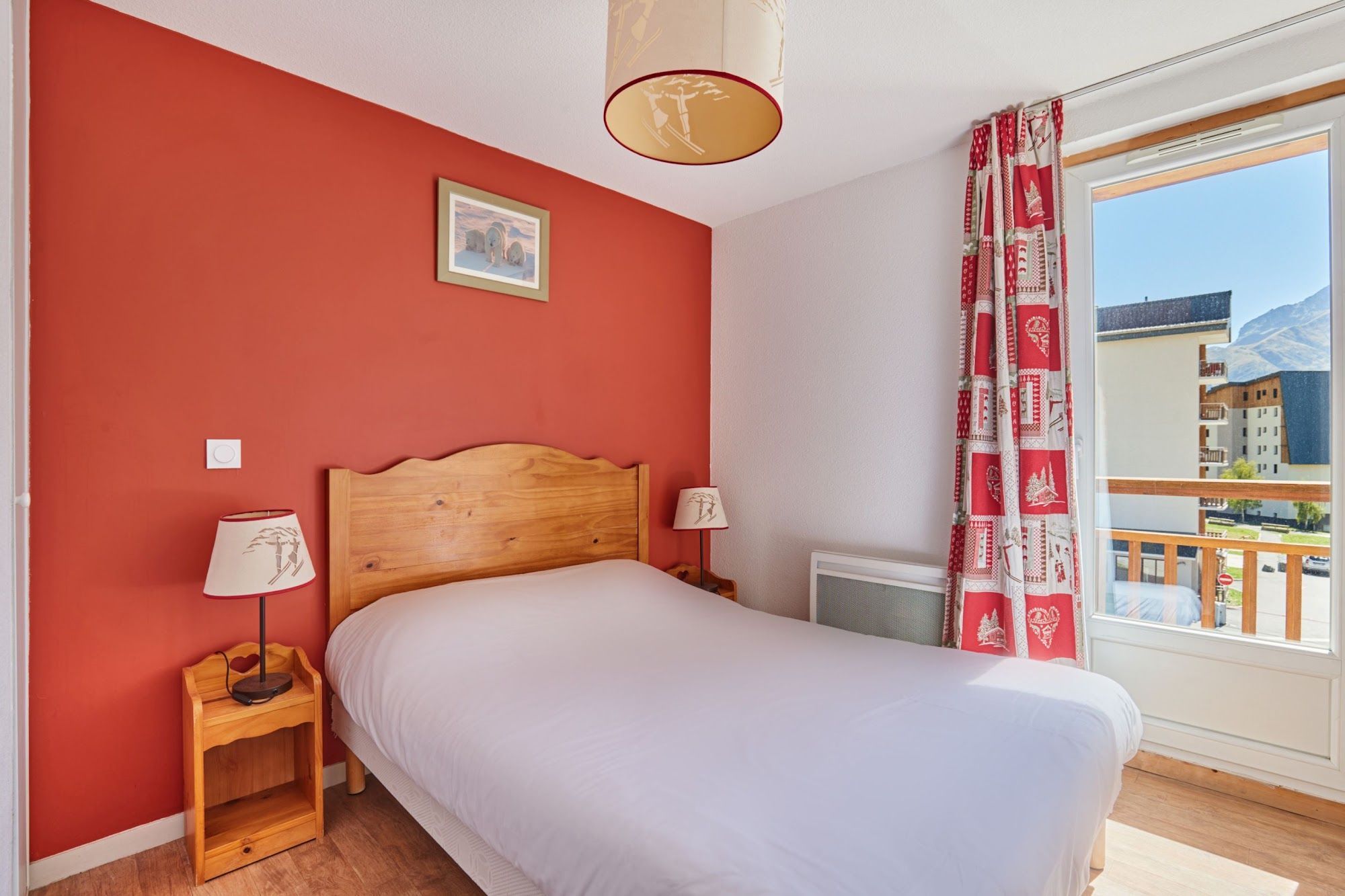 Un dormitorio con una cama de madera, una mesita de noche con una lámpara, un radiador y una ventana que ofrece vistas a edificios y montañas.