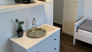 Habitación de hotel moderna con televisión de pantalla plana sobre una estantería blanca, una planta pequeña en maceta, un atril con una botella de agua y vasos, una mesita de noche blanca con cajones y una cama con almohada gris.