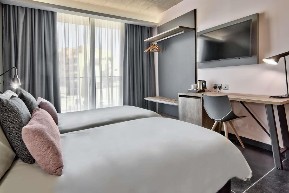 A modern hotel room with two beds, a desk, a chair, a TV, and a window with curtains.
