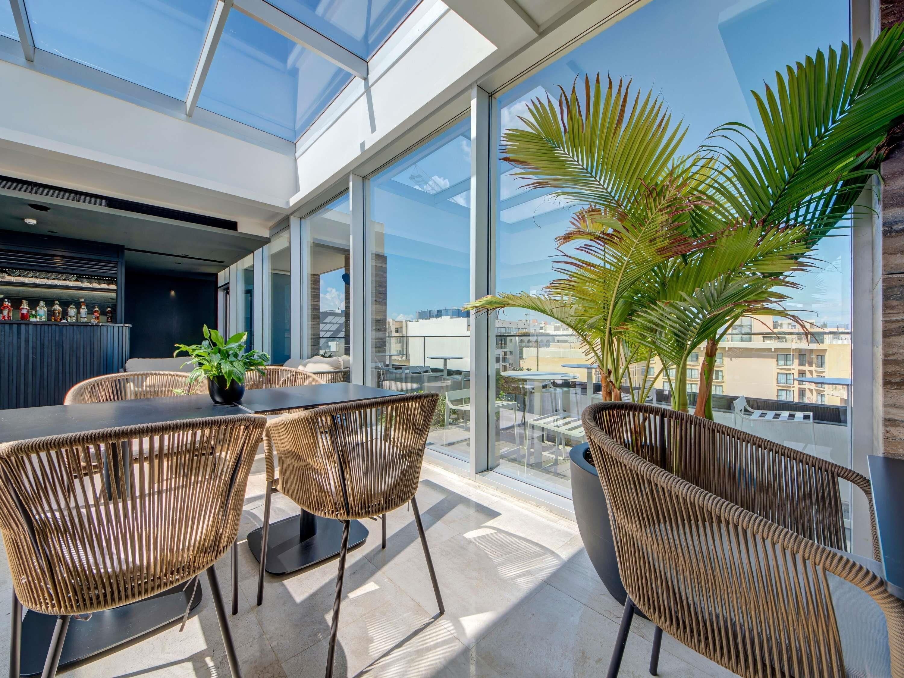 A modern interior with a dining area featuring wicker chairs and a large window overlooking a cityscape.