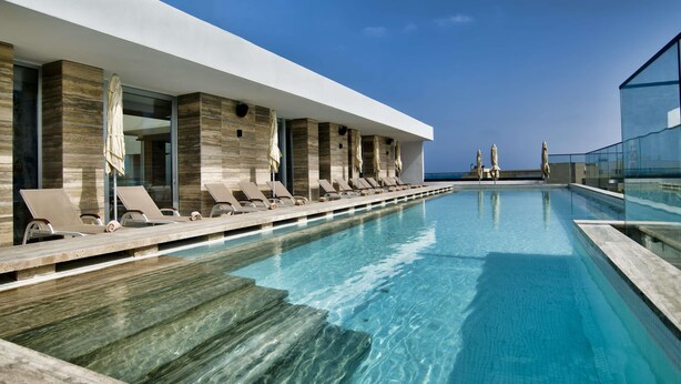 A modern outdoor pool area with lounge chairs and umbrellas, a clear blue sky, and a view of the ocean. - Number 11 St Julian's Malta - Handwritten Collection (St. Julian's)