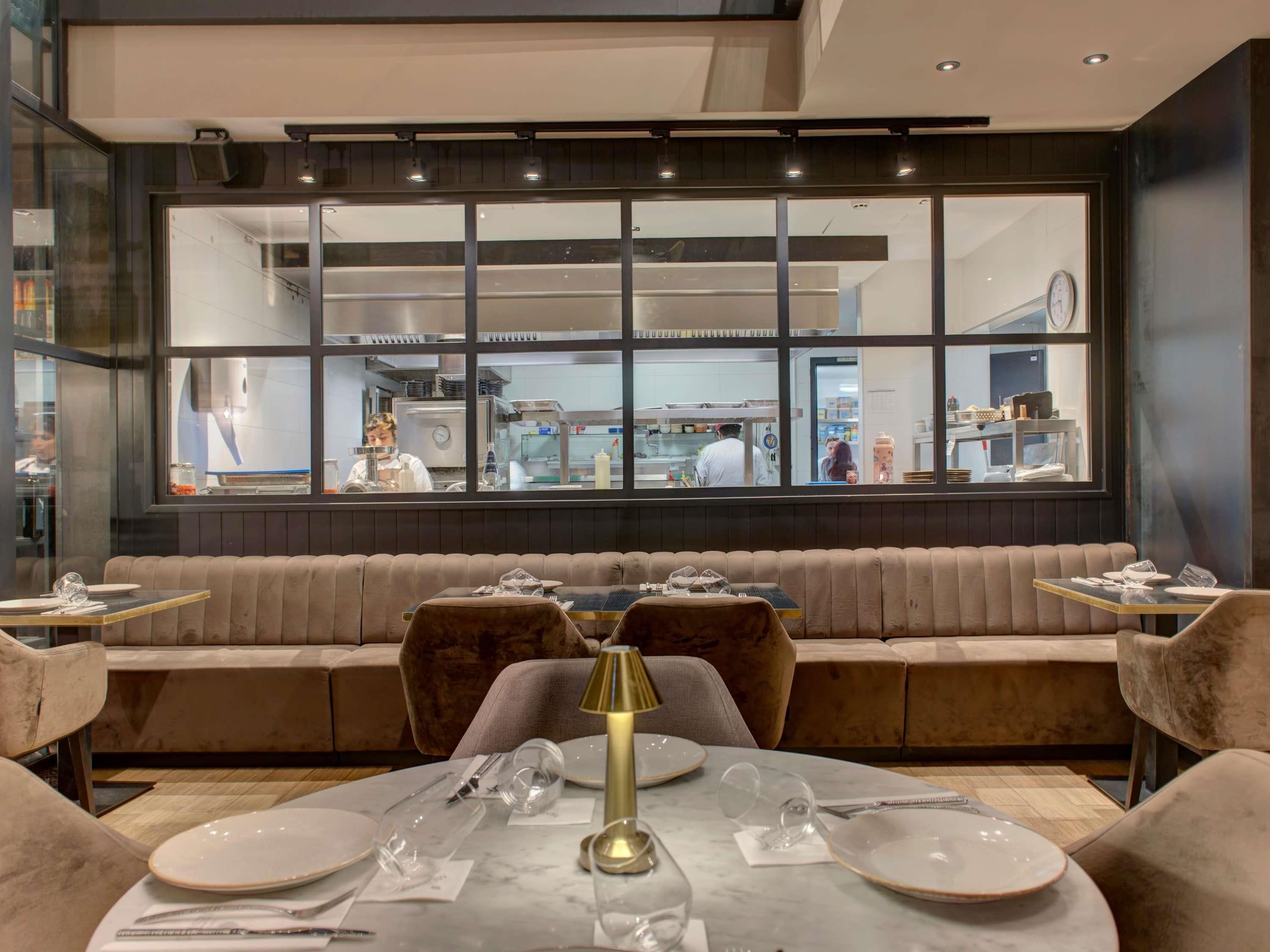 En restaurant med moderne indretning, der byder på et stort vindue med udsigt til køkkenet, et siddeområde med polstrede bænke og et marmorbord med dækket borde.