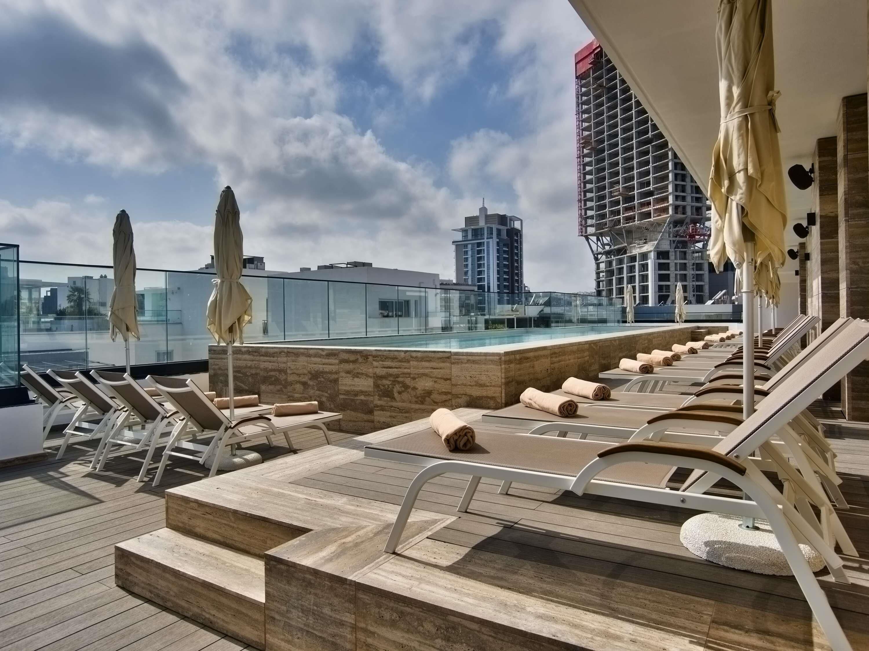 A rooftop pool area with lounge chairs and umbrellas, a glass balustrade, and a view of the city skyline.