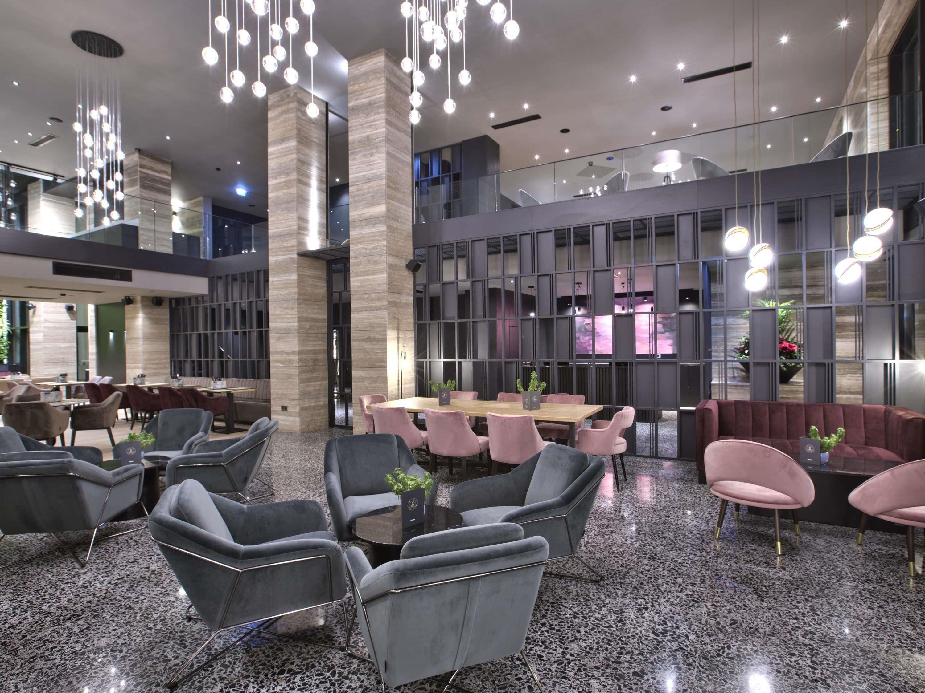 A modern hotel lobby with a central seating area, a high ceiling with hanging lights, and a large window providing a view of the interior.