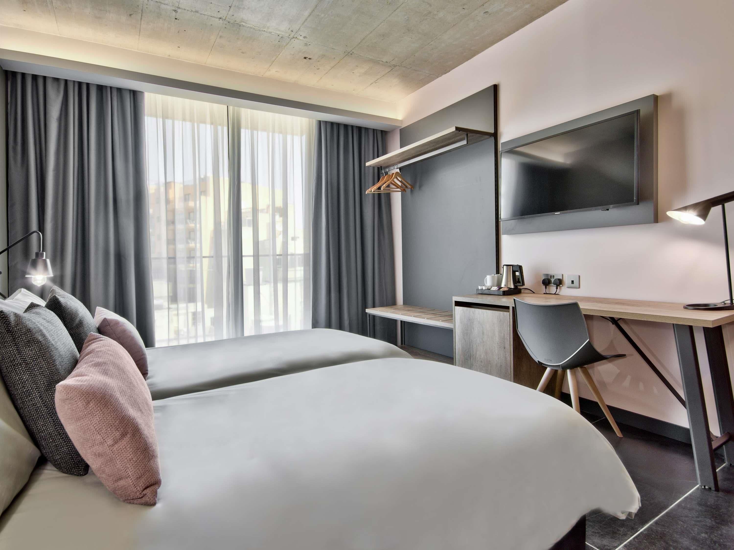 A modern hotel room with two beds, a desk, a chair, a TV, and a window with curtains.
