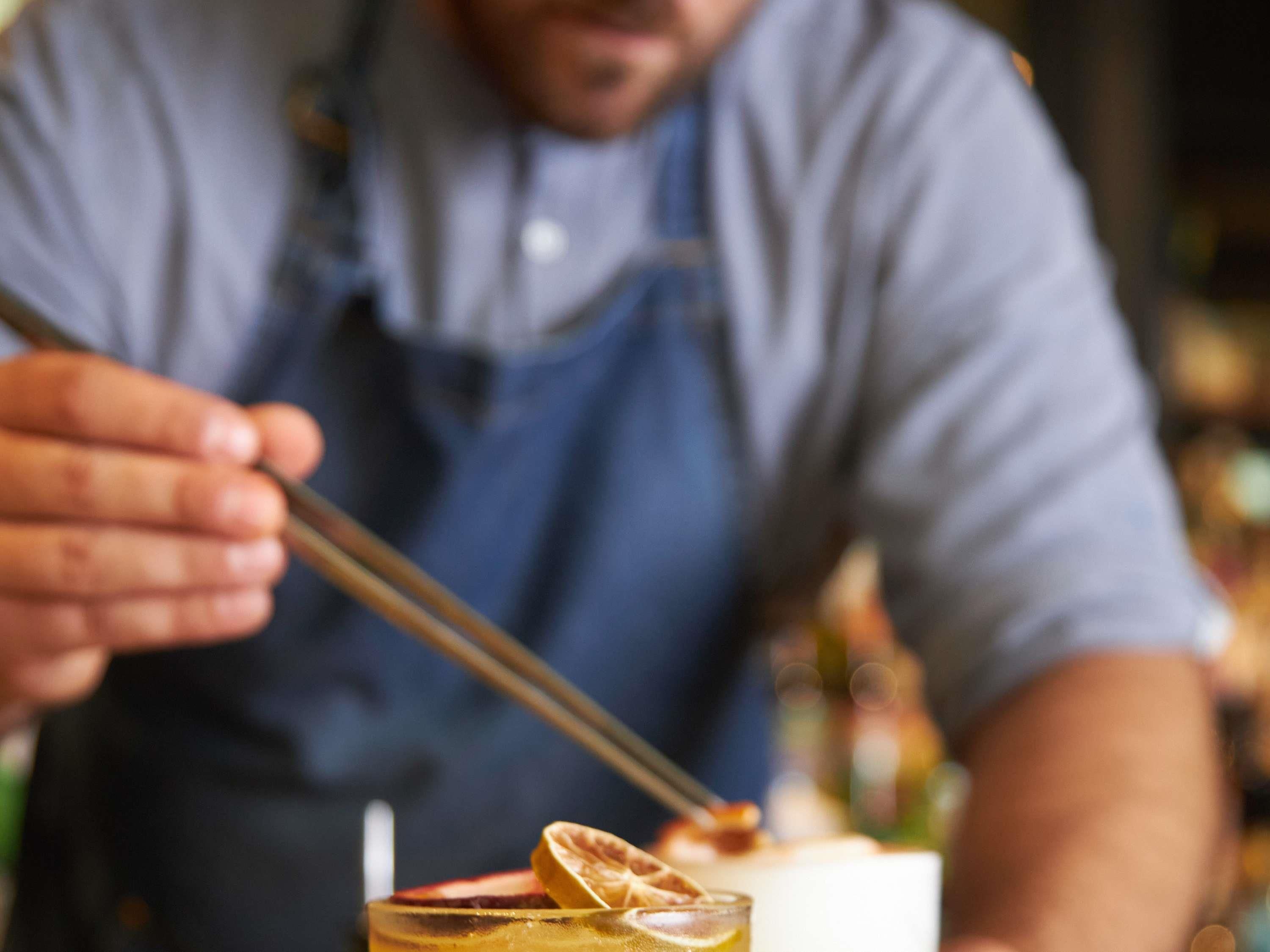 A person in a blue apron holding a pair of tongs, likely preparing a drink.