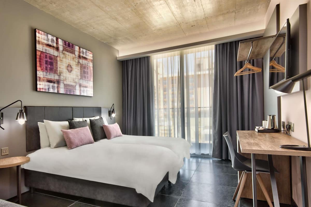 A modern hotel room with a large bed, a desk, and a view of the city through the window.