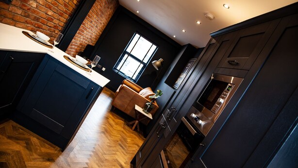 A modern kitchen with dark cabinetry, a herringbone wood floor, and a brick wall. - Barber Shop Flat (Stockport)