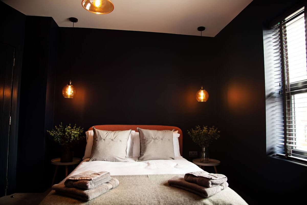 Ein schlafgemütliches Schlafzimmer in dunklen Tönen mit einem großen Bett, zwei Nachttischen inklusive Lampen und einem Fenster mit Jalousien.