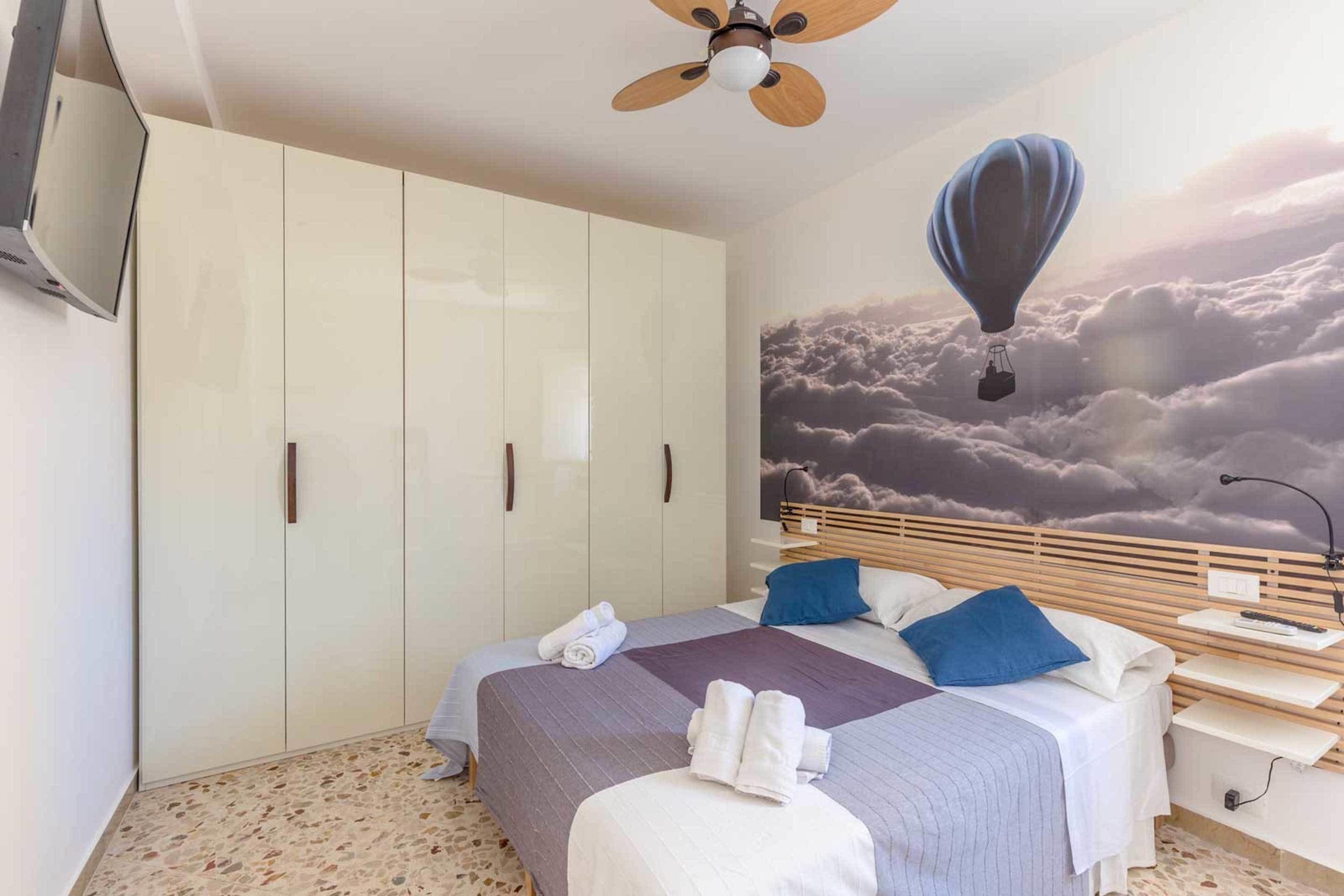 Une chambre à coucher comprenant un lit, une armoire, un ventilateur de plafond, un téléviseur fixé au mur et une fresque représentant une montgolfière.