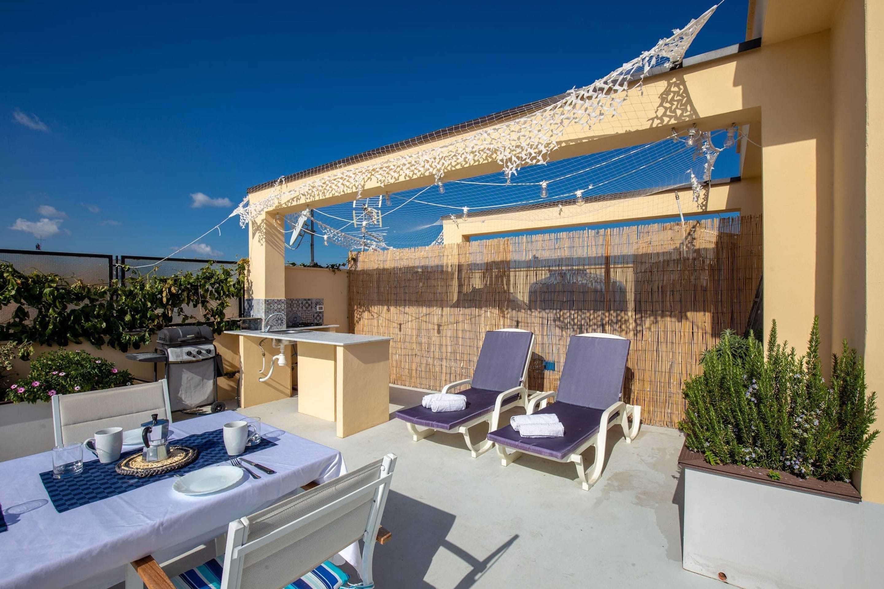 Patio esterno con tavolo apparecchiato per due persone, due sdraio, un barbecue e un piccolo angolo bar.