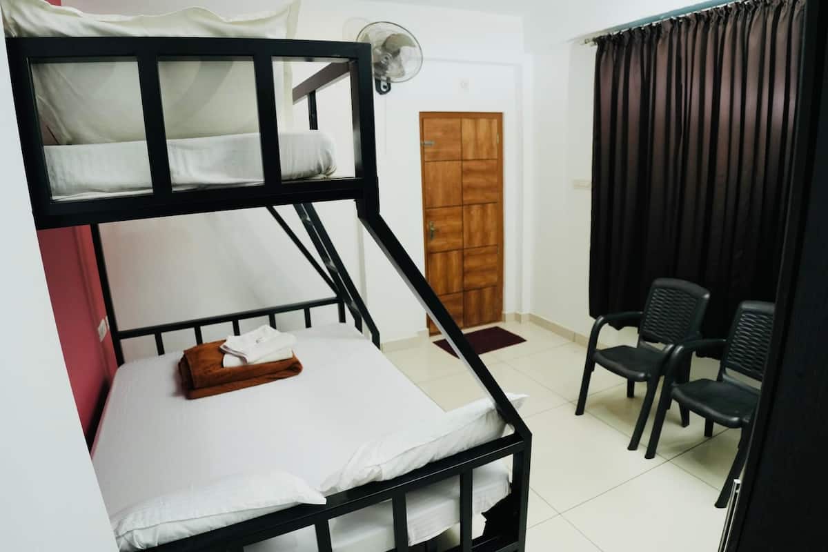 A bunk bed with a white mattress, a wooden side table, and a fan. There are two black chairs and a wooden door in the background.