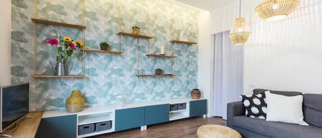 A modern living room with a patterned wallpaper, wooden shelves, a grey sofa, and a woven ottoman.