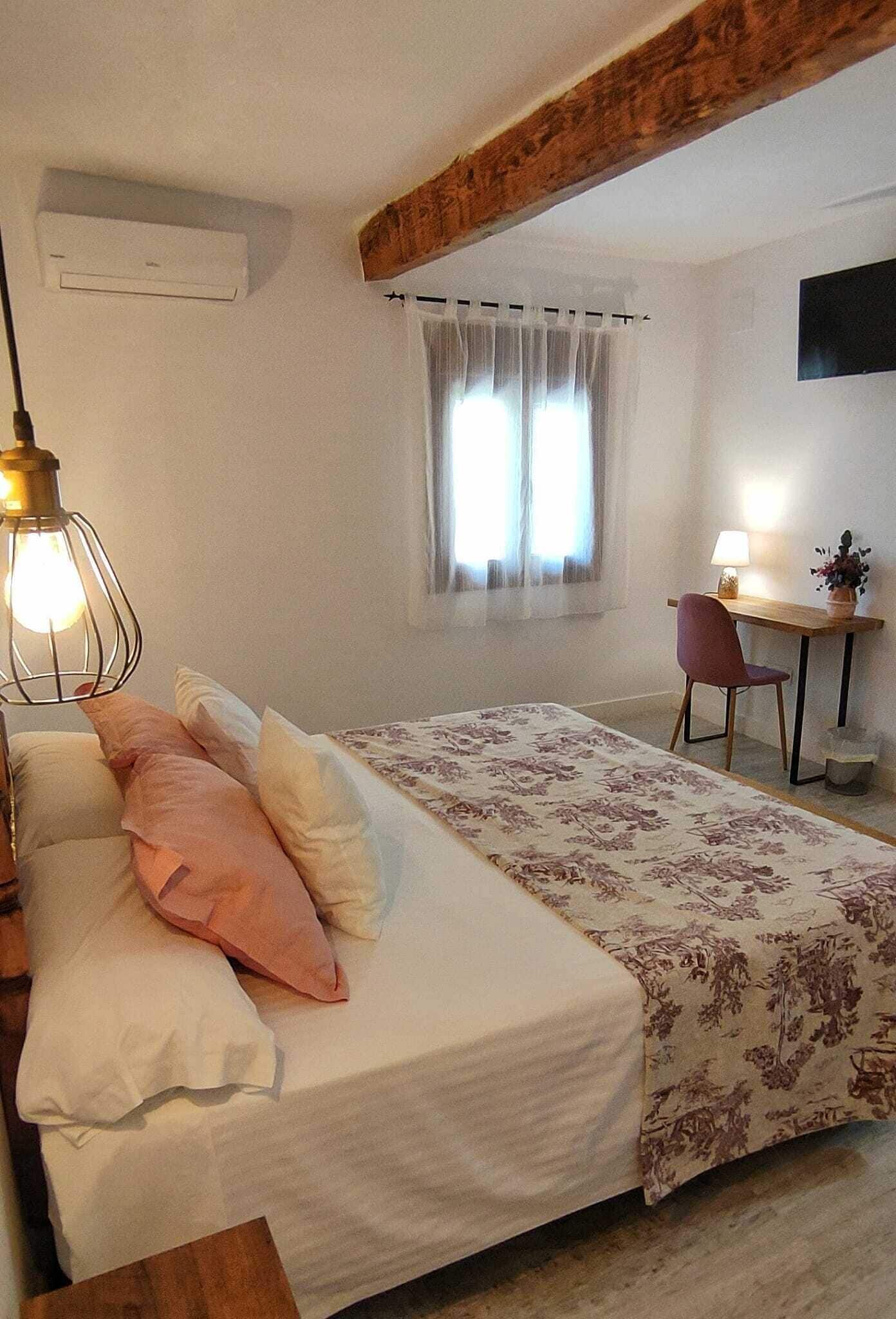 A bedroom with a bed, a desk, and a chair. There is a window with curtains, a lamp, and an air conditioning unit on the wall.