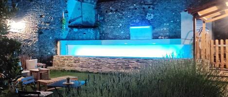 Un edificio de piedra con una torre, un área de piscina con iluminación azul, asientos al aire libre y un muro de piedra.