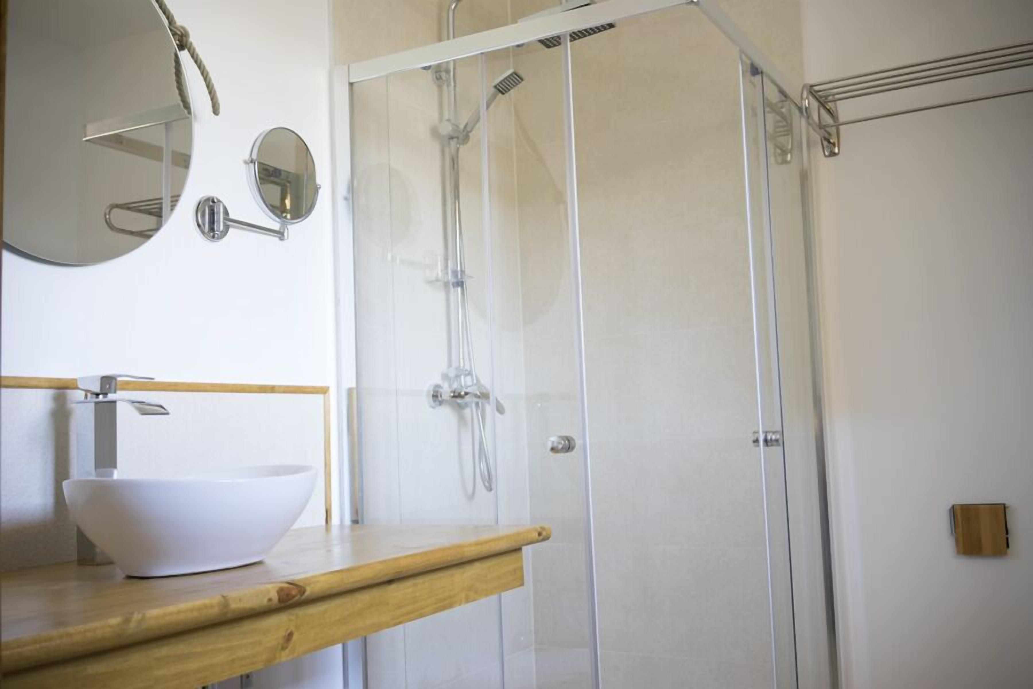 Un baño con tocador de madera, lavamanos blanco, espejo redondo y cabina de ducha con puerta de vidrio.