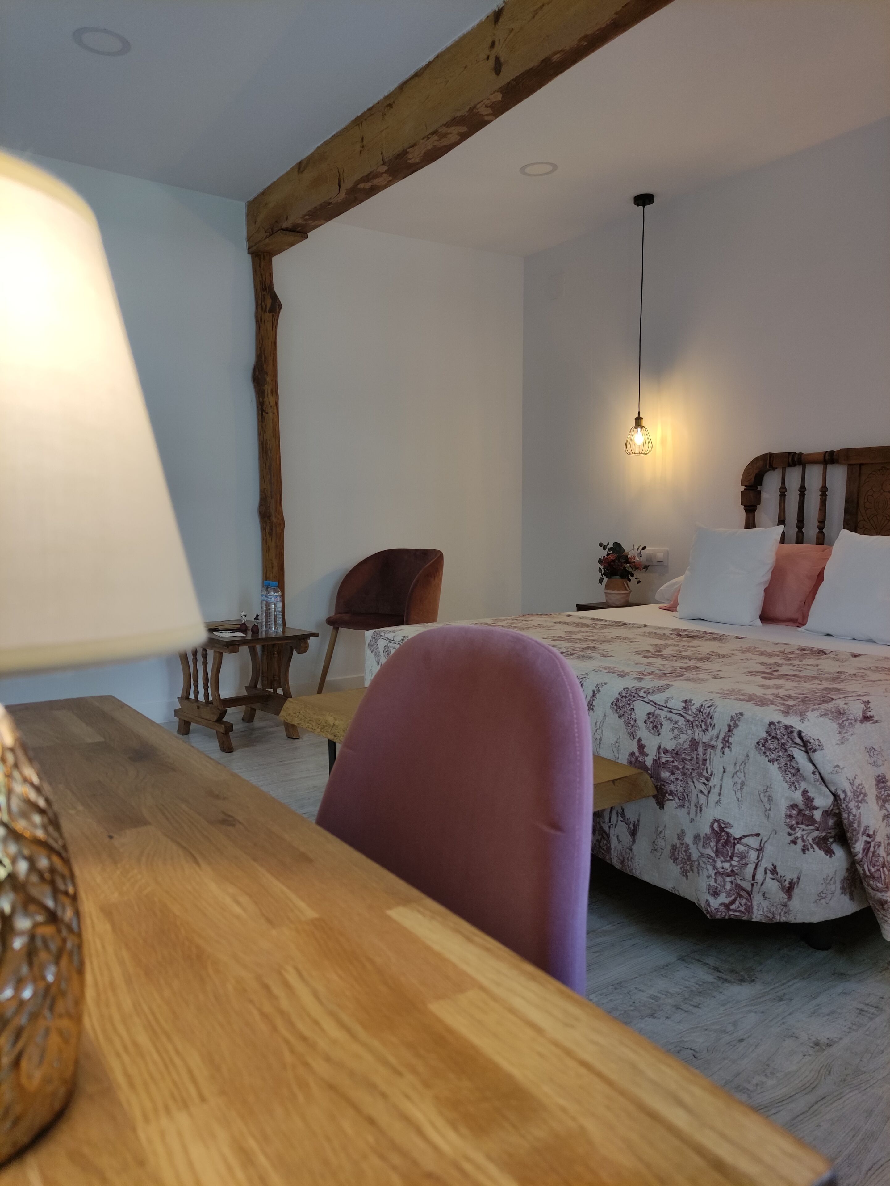 A bedroom with a wooden ceiling, a bed with floral bedding, a bedside table, a chair, and a lamp.