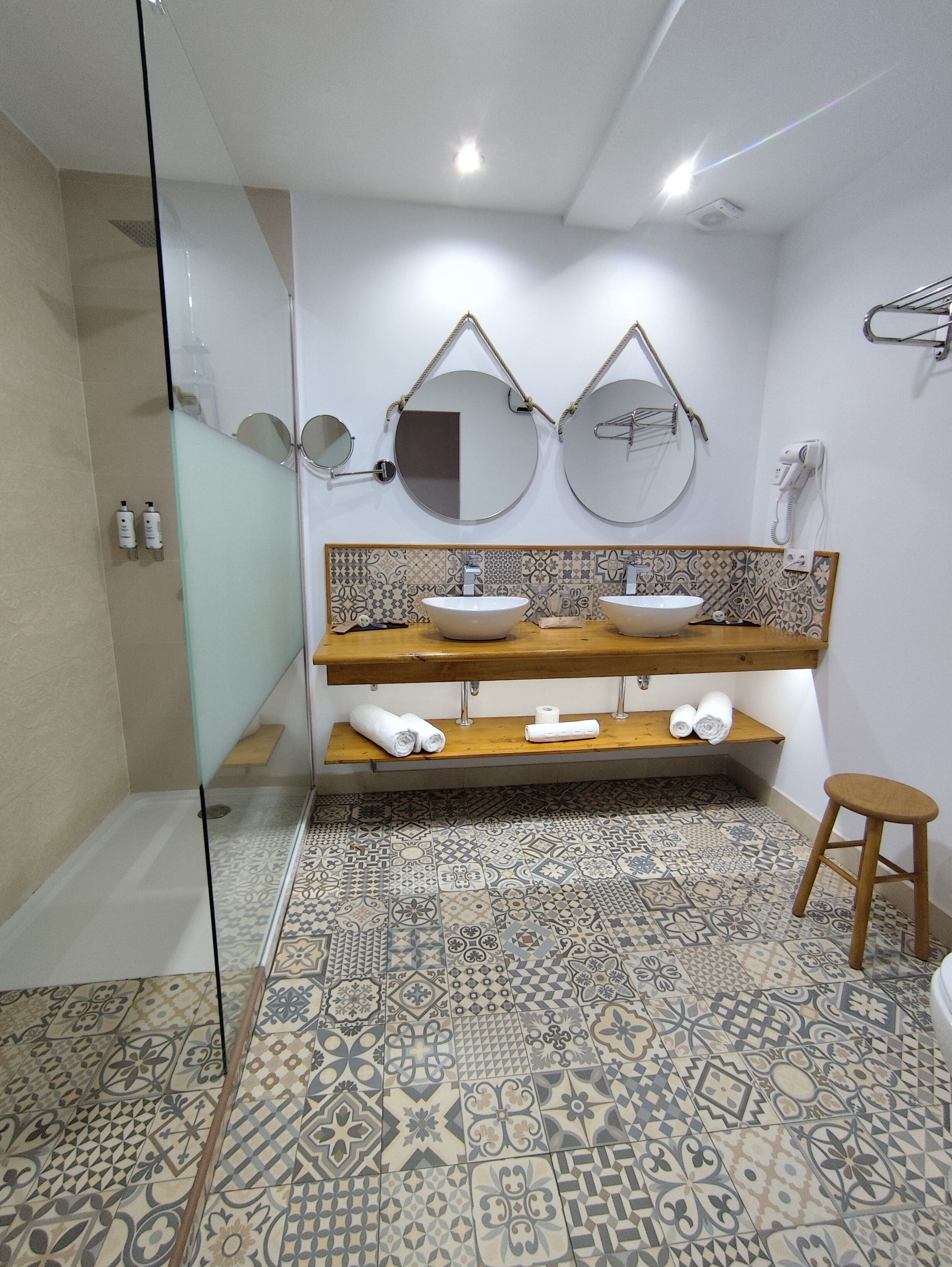 A bathroom with two sinks, a shower, and a toilet. There are two mirrors above the sinks and a hairdryer on the wall. The floor has patterned tiles.