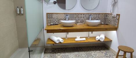 Un baño con dos lavamanos, una ducha y un inodoro. Hay dos espejos sobre los lavamanos y un secador de pelo en la pared. El suelo tiene baldosas con diseños.