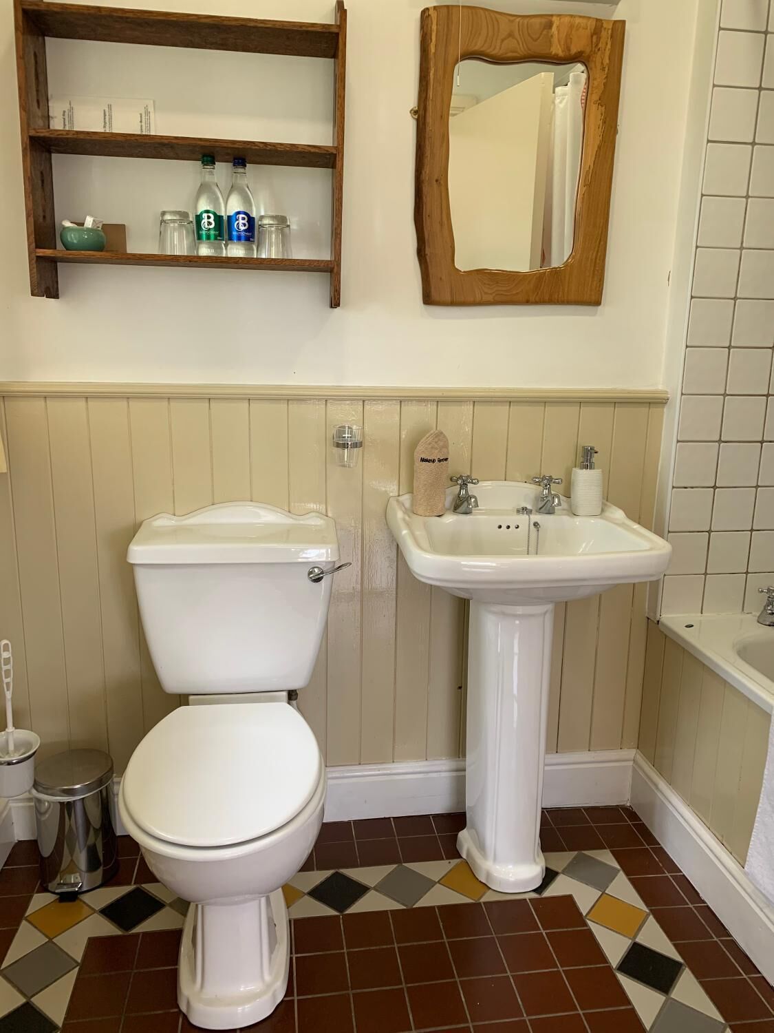 Et badeværelse med toilet, vask og spejl. Der er en hylde med toiletartikler og en skraldespand.