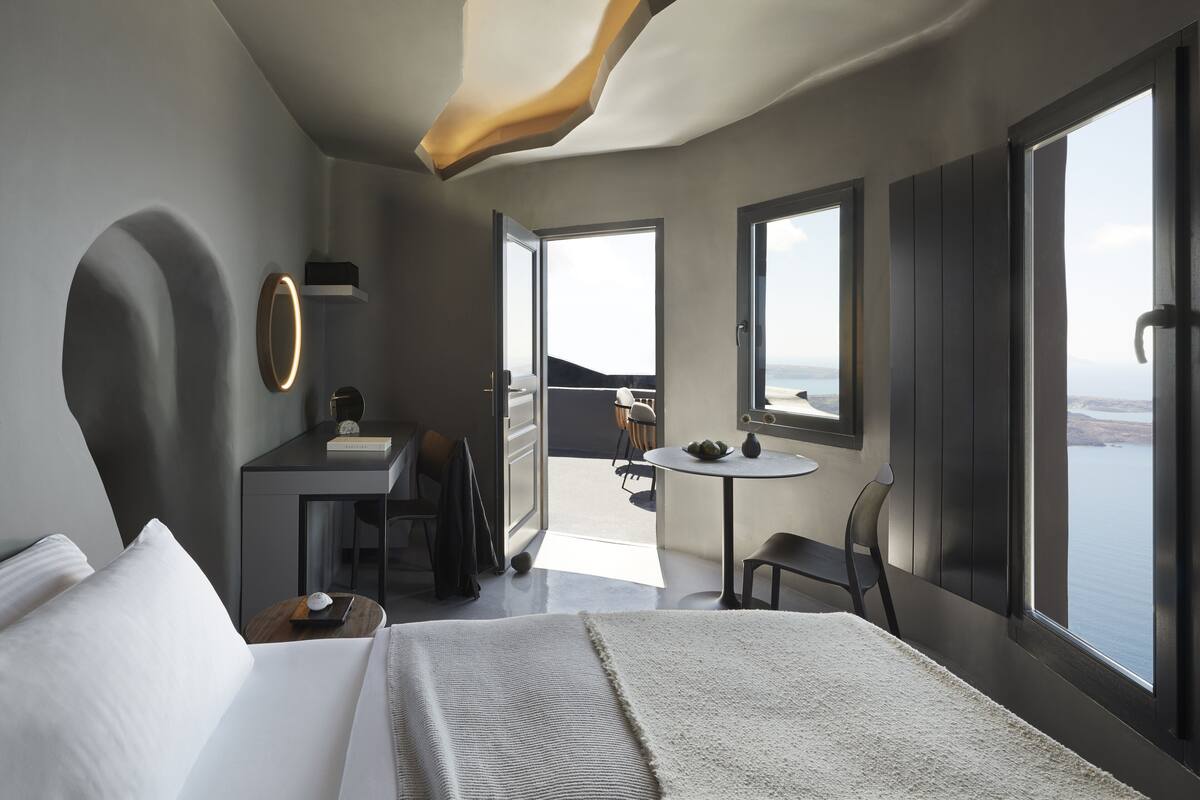 A modern hotel room with a large bed, a desk with a chair, a small table with a bowl, and a view of the sea through large windows.