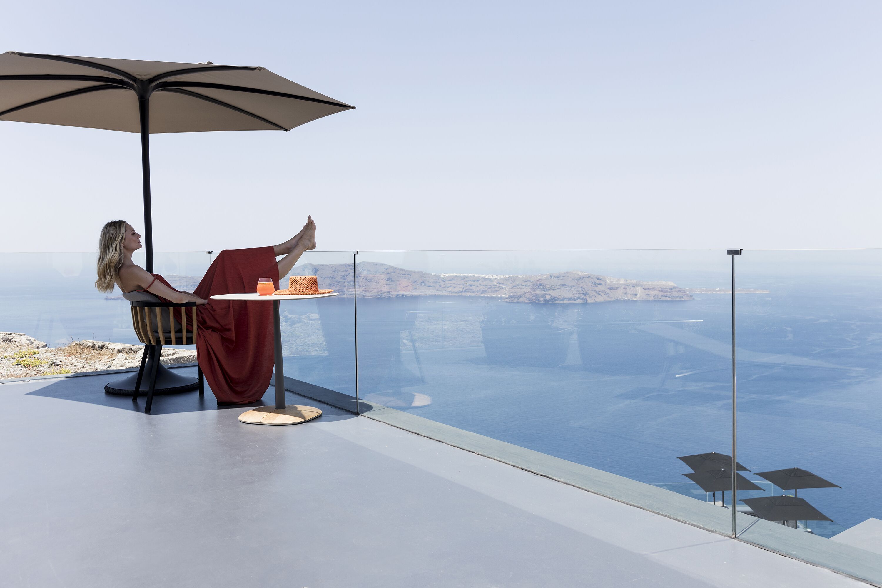 Eine Person entspannt sich auf einem Balkon mit Tisch und Sonnenschirm, mit Blick auf eine malerische Seelandschaft und das entfernte Land.