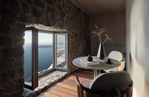 A room with a stone wall, a round table with a book and vase, and a view of the sea. - Kivotos Santorini - Preferred Hotels & Resorts (Santorini)