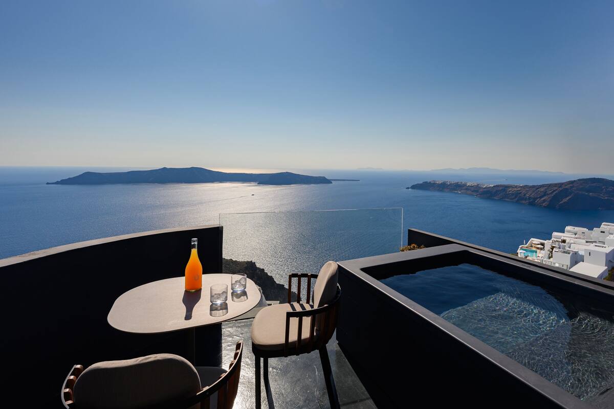 A modern outdoor seating area with a table, chairs, and a hot tub overlooking a body of water and distant islands.