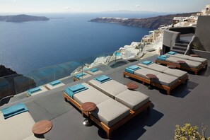 A rooftop lounge with sunbeds and round tables, overlooking a coastal sea and mountainous landscape. - Kivotos Santorini - Preferred Hotels & Resorts (Santorini)