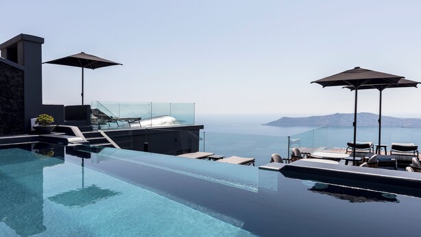 Una piscina infinita con vistas despejadas al mar y a las montañas, con mobiliario exterior y sombrillas.