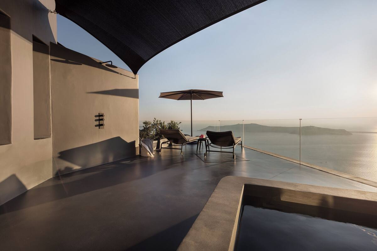 A modern outdoor lounge area with a view of the sea, featuring a pool, lounge chairs, and an umbrella.