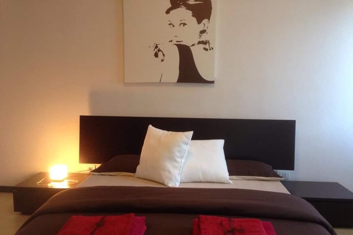 A hotel room with a bed, two pillows, a bedside table with a lamp, and a wall-mounted artwork.