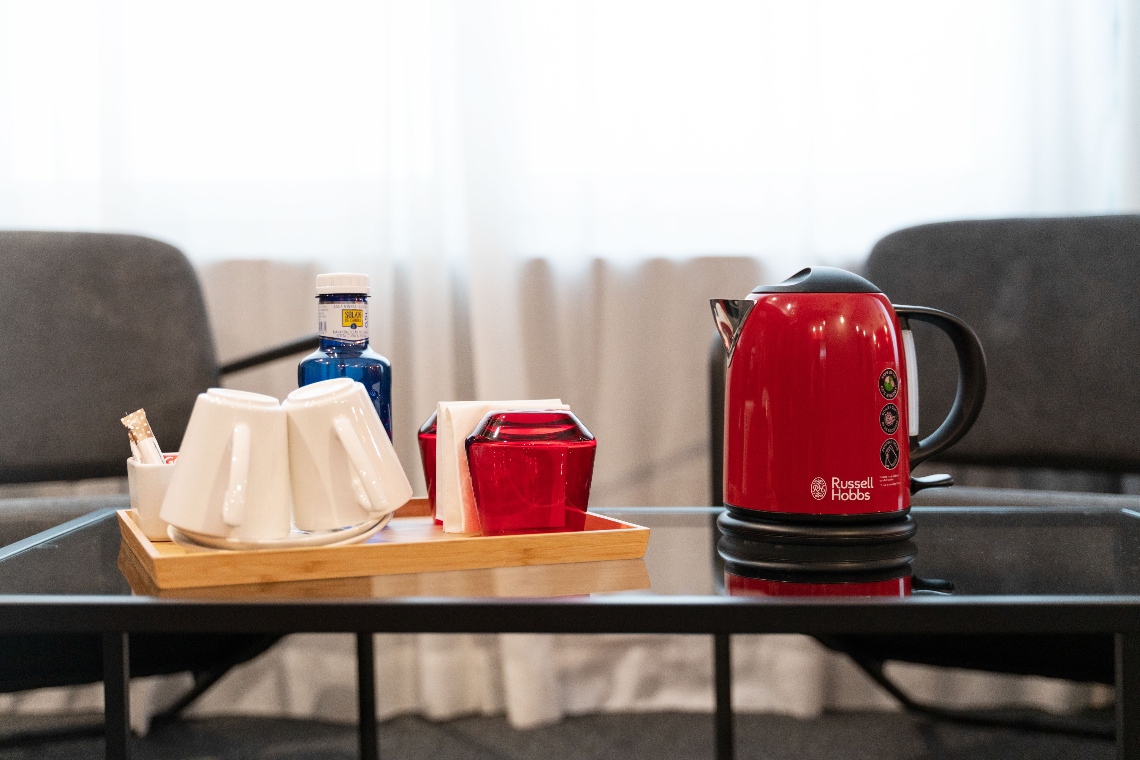 A table with a red kettle, white and red cups, and a blue bottle on a tray.