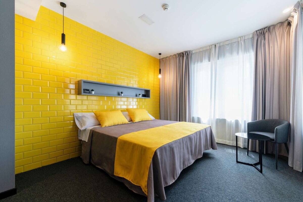 Ein Hotelzimmer mit einer gelben Ziegelwand, einem Bett mit einer grau-gelben Tagesdecke, einem grauen Sessel und einem kleinen Tisch.