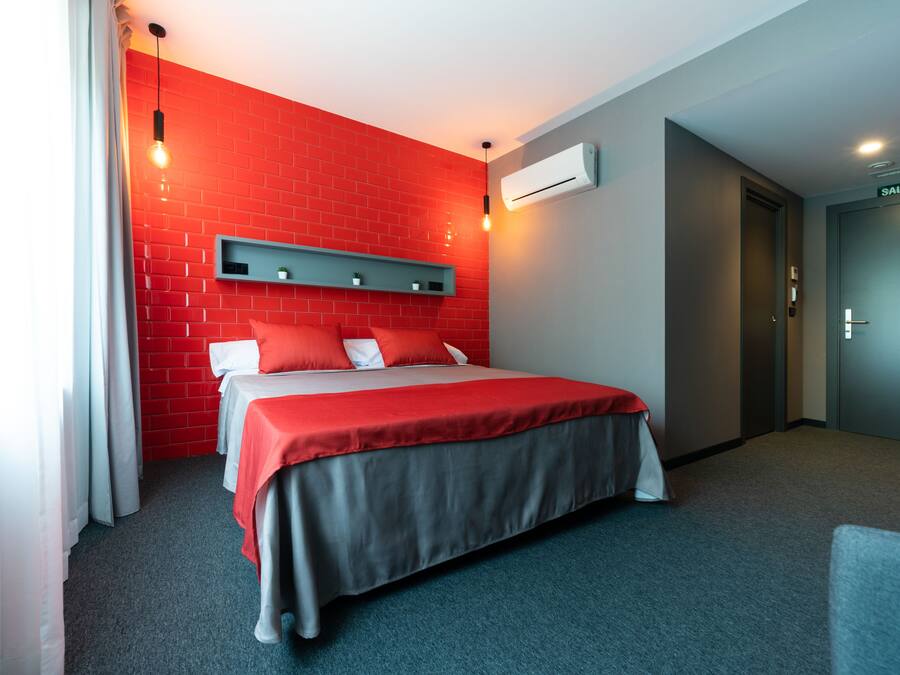 A hotel room with a red brick wall, a bed with red and grey bedding, a grey headboard, a small shelf with plants, and an air conditioning unit on the wall.