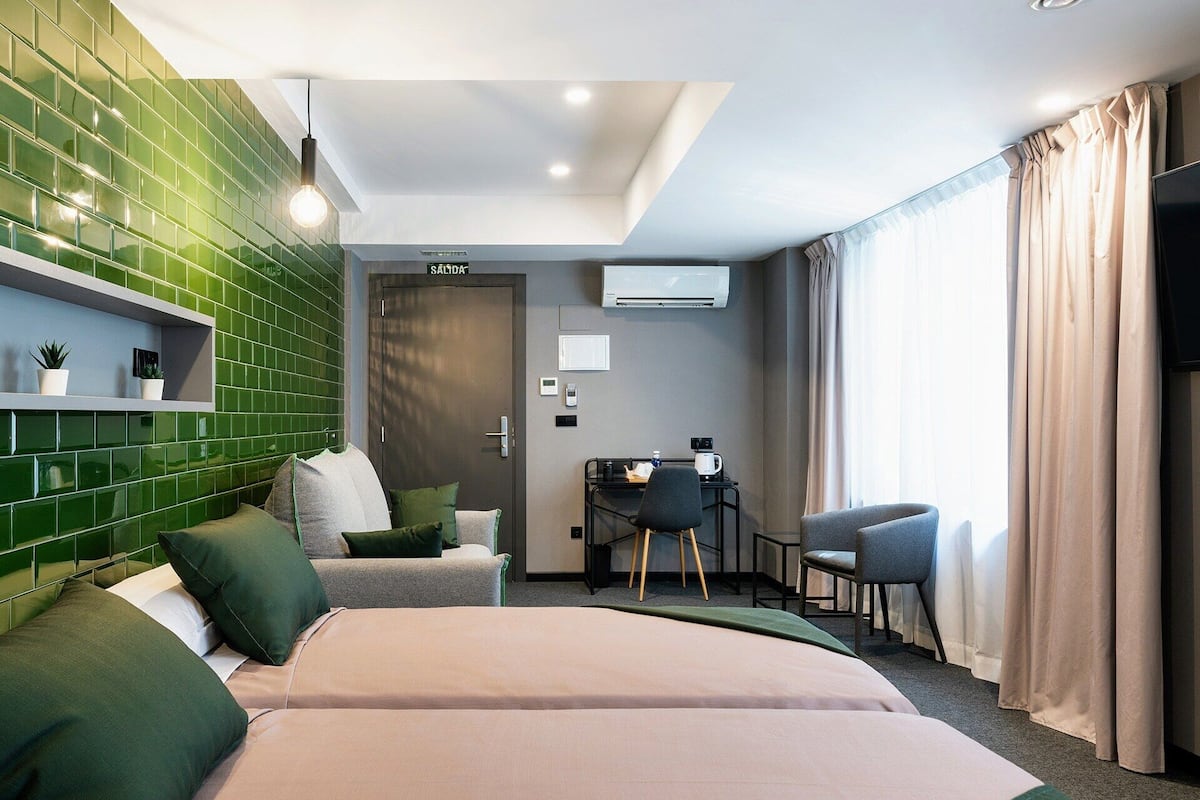 Ein Hotelzimmer mit zwei Betten, einem Schreibtisch mit Stuhl, einem kleinen Tisch mit Kaffeemaschine und einer grünen Wand mit Fliesen.