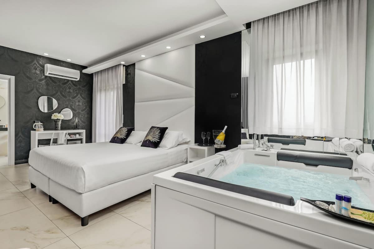 A modern bedroom with a large bed, a separate sitting area with a small table, and a bathtub with amenities.