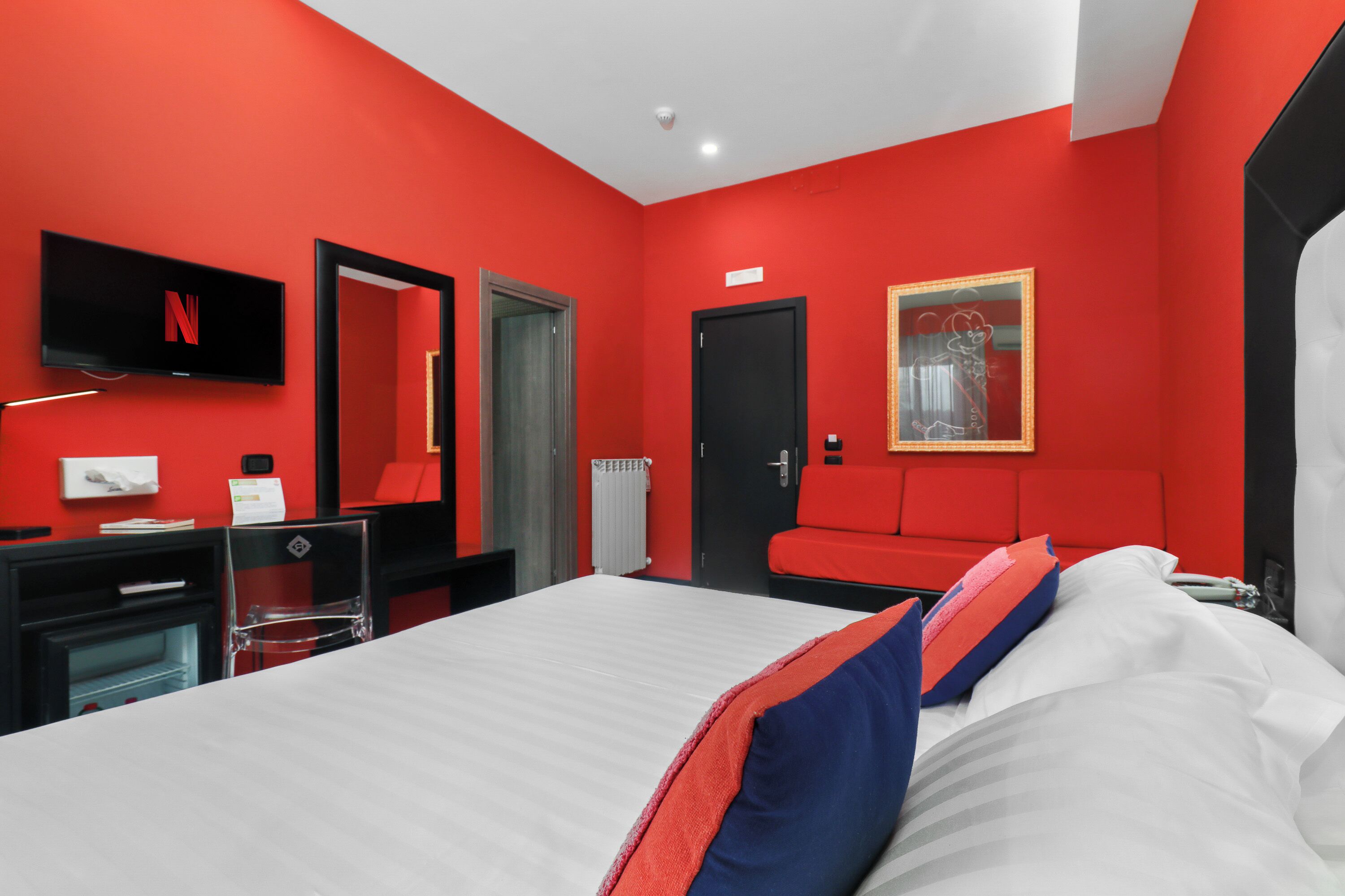 A modern hotel room with a red accent wall, a flat-screen TV, a bed with white and red bedding, a black door, and a mirror with a decorative frame.