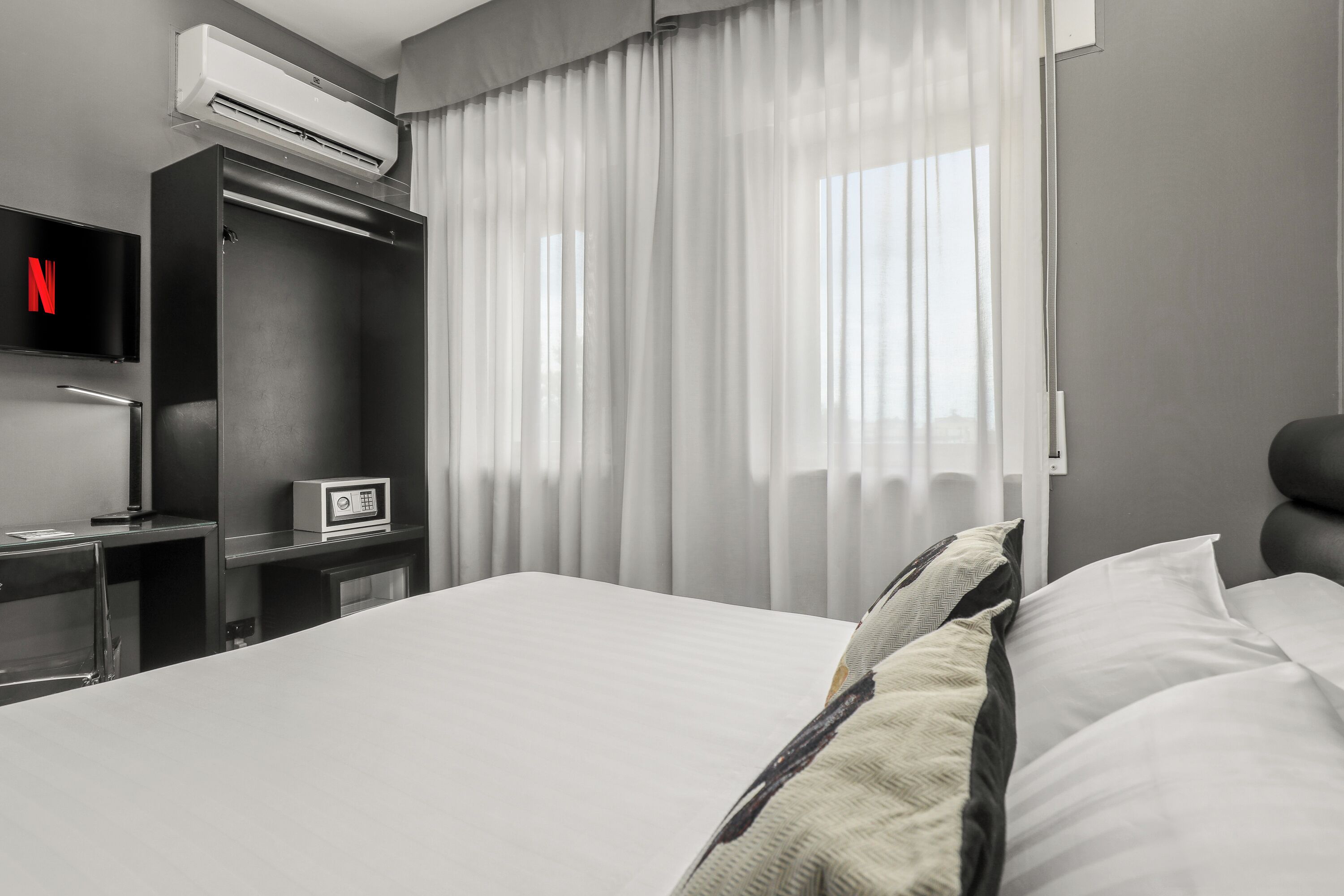 Una habitación de hotel moderna con una cama grande, un televisor de pantalla plana, un minibar y una ventana con cortinas translúcidas.