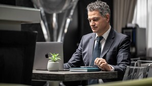 Un hombre trajeado sentado en un escritorio con una computadora portátil, una planta y un cuaderno.