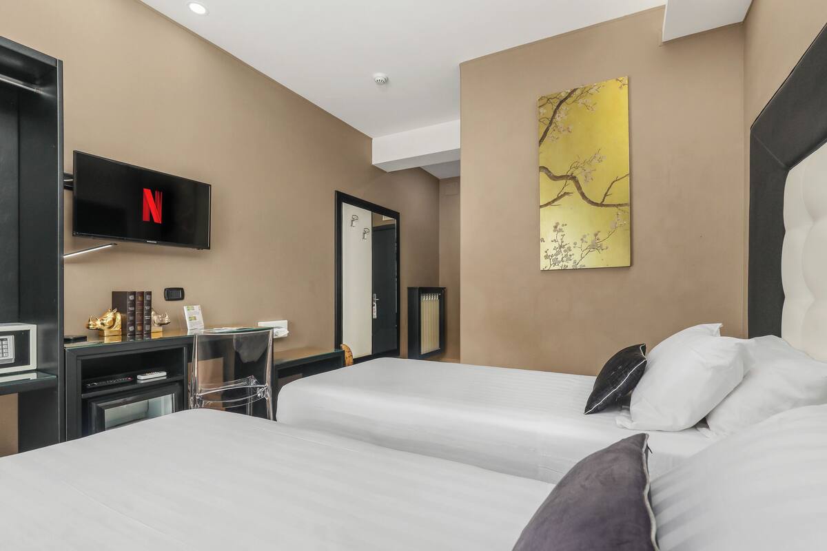 A hotel room with two beds, a desk with a TV displaying the Netflix logo, a large mirror, and a decorative wall piece.