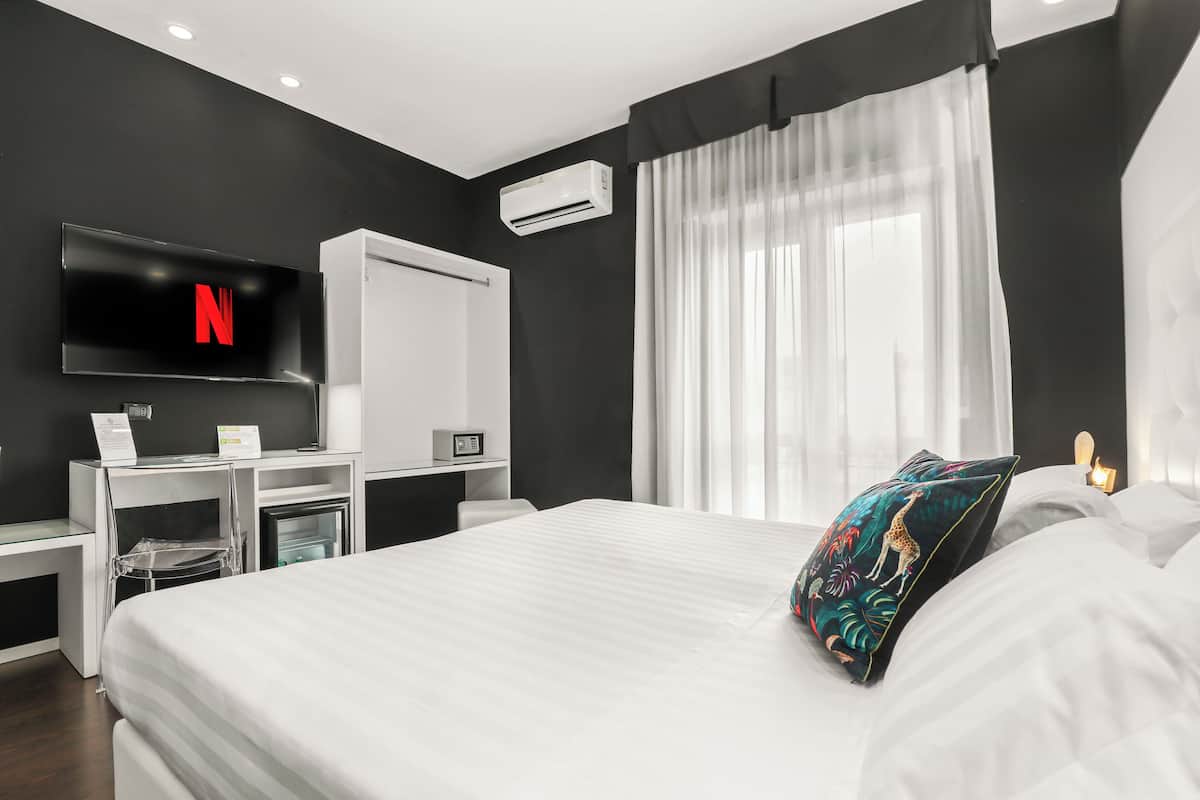 A modern hotel room with a large bed, a TV mounted on the wall, a desk with a computer, and a window with sheer curtains.