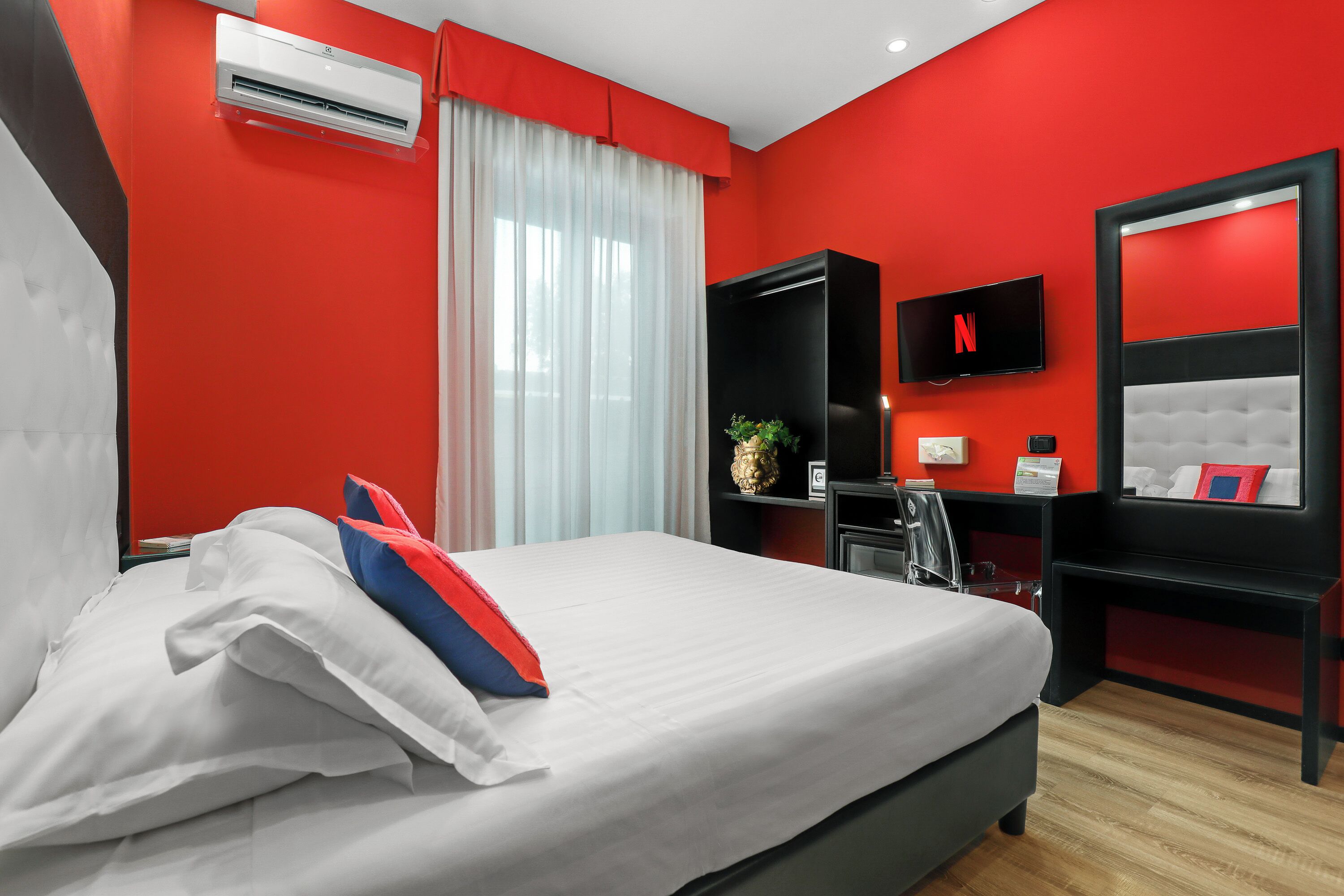 Habitación de hotel con pared roja, cama blanca, televisor negro y un espejo grande.
