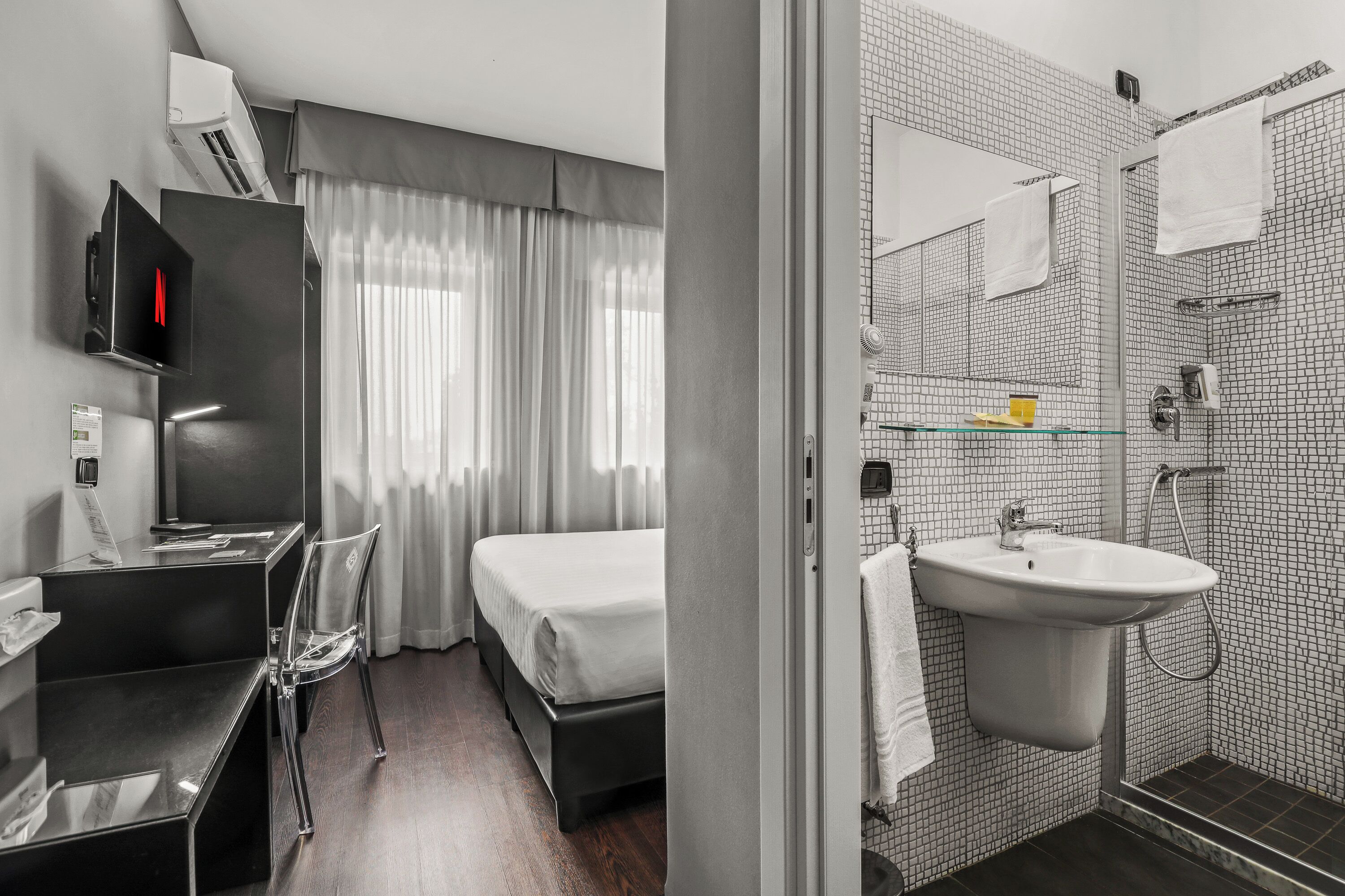 Habitación de hotel con cama, escritorio con silla, televisión y baño con lavamanos y ducha.
