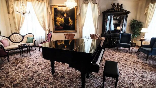 Una sala imponente con un piano de cola negro, un cuadro grande, una araña y muebles clásicos.