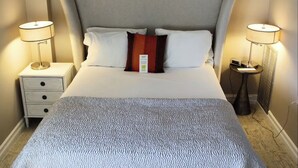 Una cama bien tendida con un cabecero gris, dos lámparas de noche y una mesita de noche con una radio.
