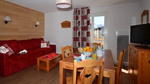 A cozy living room with a red sofa, wooden dining table, and chairs. A television is on a wooden cabinet, and a window with a view of buildings is visible. - Appartement abordable | Accès piscine intérieure (Les Deux Alpes)