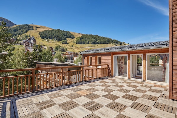 A wooden deck with a checkered floor pattern, a wooden railing, and a view of a mountainous landscape.