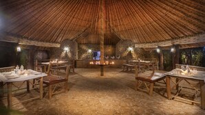 Un comedor interior con suelo de piedra, mesas y sillas de madera, y una chimenea central con velas.